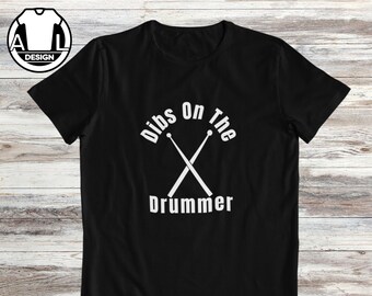 Puede incluir: Camiseta negra con el texto blanco "Dibs on the Drummer" y baquetas cruzadas. La camiseta tiene un logotipo redondo en la esquina superior izquierda.