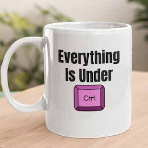 Könnte beinhalten: Weiße Keramiktasse mit einer schwarzen Aufschrift "Everything is Under Ctrl" mit einer lilafarbenen Knopfgrafik.