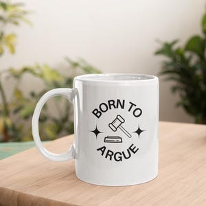 Puede incluir: Taza de café de cerámica blanca con un gráfico negro que dice "Born to Argue" con un martillo y ondas sonoras.
