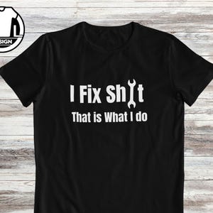 Op de afbeelding: Een zwart t-shirt met korte mouwen en witte tekst I Fix Sh*t That is What I do, waarbij de letter I in het woord Shit is vervangen door een witte afbeelding van een steeksleutel. Het ontwerp staat centraal op de voorzijde.