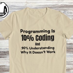 Op de afbeelding: Een beige t-shirt met korte mouwen en zwarte tekst: Programming Is 10% Coding And 90% Understanding Why It Doesn't Work. Het t-shirt heeft een ronde hals en een klein rond logo in de linkerbovenhoek met de letters A en L.