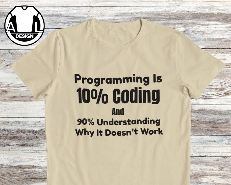 Op de afbeelding: Een beige t-shirt met korte mouwen en zwarte tekst: Programming Is 10% Coding And 90% Understanding Why It Doesn't Work. Het t-shirt heeft een ronde hals en een klein rond logo in de linkerbovenhoek met de letters A en L.