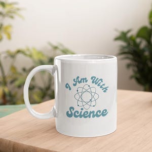Könnte beinhalten: Weiße Keramiktasse mit einer blauen Grafik, auf der "I Am With Science" und ein stilisiertes Atom-Symbol stehen.