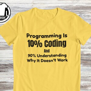 Op de afbeelding: Een felgeel T-shirt met korte mouwen en zwarte tekst: Programming Is 10% Coding And 90% Understanding Why It Doesn't Work. Het shirt heeft een ronde hals en ligt plat op een lichtgekleurde houten ondergrond.