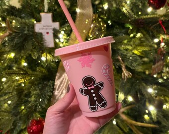 May include: A pink plastic tumbler with a matching lid and straw. The tumbler is decorated with a gingerbread man, snowflakes, and candy cane graphics. The tumbler is held in front of a Christmas tree.