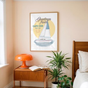May include: A framed wall art print featuring a sailboat illustration with a yellow circle background. The text reads Greetings from CAPE COD YACHT CLUB. The sailboat has white sails with blue accents and a white hull with red and blue horizontal stripes.