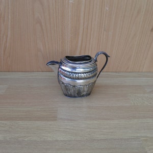 May include: A silver creamer with a decorative design around the top and bottom. The creamer has a handle and a spout.