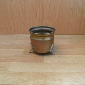 May include: A small, brass planter with three decorative feet. The planter is empty and has a shiny, gold finish.