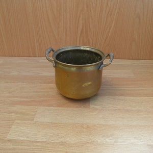 May include: A small, brass planter with two handles. The planter is empty and has a worn, aged look.