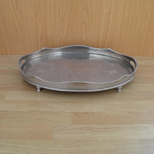 May include: A silver oval serving tray with a decorative border and three feet. The tray has a slightly raised edge and a smooth surface.