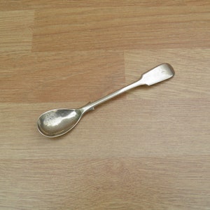 May include: A silver teaspoon with a rounded bowl and a plain handle. The spoon is lying on a light brown wooden surface.