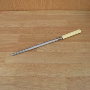 May include: A silver steel knife sharpener with a white plastic handle. The sharpener is lying on a light brown wooden surface.