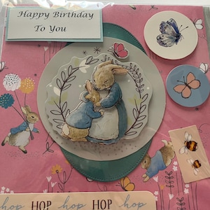 May include: A handmade birthday card featuring illustrations of Peter Rabbit and other characters. The card has a pink floral background and includes the text "Happy Birthday To You". Decorative butterfly and bee stickers are also included.