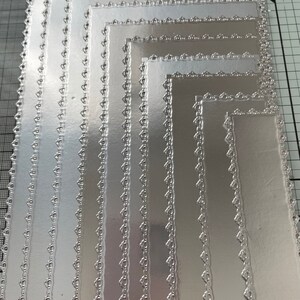 May include: A stack of silver rectangular cardstock with decorative scalloped edges. The cardstock is arranged in a nested pattern, with each piece slightly smaller than the last. The edges feature a repeating heart-shaped design.
