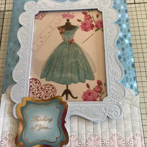 May include: A handmade greeting card with a light blue frame and a picture of a teal dress on a mannequin. The card has a decorative border and a gold-trimmed label that reads "Thinking of You..."