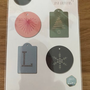 May include: Sizzix Thinlits die set featuring four tag designs: a pink burst, a green Christmas tree, a blue letter "L", and a gray snowflake. The package includes the text "JOSH GRIFFITHS" and "6 DIES".