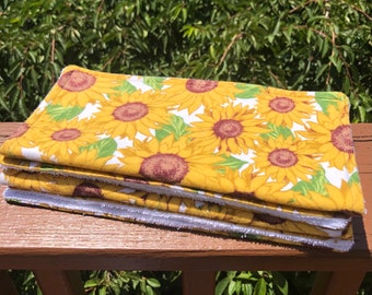May include: A stack of four yellow dish towels with a sunflower print. The towels have white borders and are folded neatly.