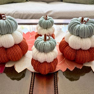 May include: Three stacked pumpkin decorations made of ribbed knit fabric. Each stack consists of a light blue top, a white middle, and a burnt orange bottom pumpkin. Each pumpkin features a small brown stem at the center of the top section.