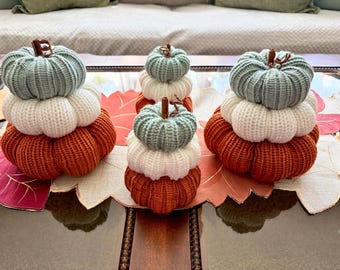 May include: Three stacked pumpkin decorations made of ribbed knit fabric. Each stack consists of a light blue top, a white middle, and a burnt orange bottom pumpkin. Each pumpkin features a small brown stem at the center of the top section.