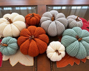 May include: A collection of eight decorative pumpkins crafted from ribbed knit fabric in shades of cream, orange, gray, and sage green. Each pumpkin features a small, brown cinnamon stick stem protruding from the center of its rounded top.