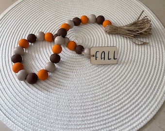 May include: A fall-themed garland made of wooden beads in brown, orange, and beige colors. The garland has a brown wooden tag with the word "FALL" written in black.