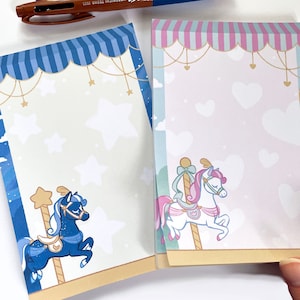 May include: Two decorative memo pads with a carousel theme. The left pad features a blue horse with stars and a crescent moon. The right pad features a pink horse with hearts and a sun.