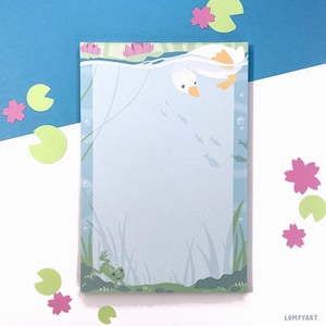 May include: A notepad with a whimsical illustration of a duck swimming underwater, surrounded by fish, aquatic plants, and a frog. The notepad has a light blue background with a border. Pink flower and green lily pad cutouts are scattered around the notepad.