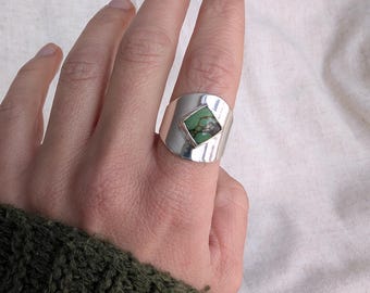 Puede incluir: Un anillo ancho de plata con una cara rectangular biselada. La cara presenta una piedra cuadrada verde con inclusiones marrones y negras. El anillo se lleva en un dedo, con una manga verde visible.