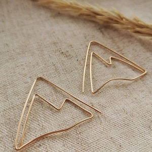 May include: A pair of gold-toned earrings shaped like abstract mountains. The earrings are made of thin wire and have a delicate, minimalist design.