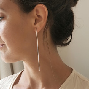 May include: A person wearing a long, thin silver earring. The earring is straight and hangs down past the person's shoulder.