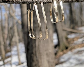 Op de afbeelding: Goudkleurige oorringen in verschillende maten hangen aan een tak. De oorbellen hebben een rechthoekige vorm met afgeronde randen. De achtergrond toont een wazige bosscène met bomen en sneeuwplekken.