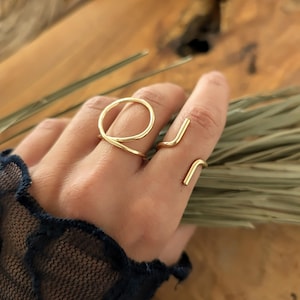May include: Two gold-colored rings are displayed on a hand. One ring features a circular design, while the other has a minimalist, angular shape. The rings are made of a shiny, metallic material, and are set against a blurred background.