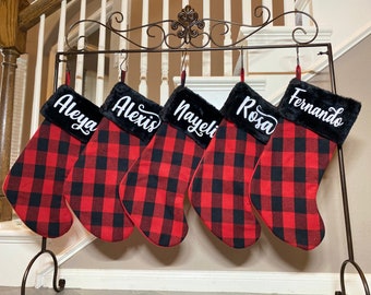 May include: Five Christmas stockings hanging on a black metal stand. Each stocking features a red and black buffalo plaid pattern with a black faux fur cuff. The names Aleya, Alexis, Nayeli, Rosa, and Fernando are printed in white script on the cuffs.