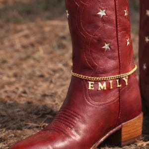 Op de afbeelding: Rode leren cowboylaarzen met steruitsparingen en een gouden enkelband met de naam "EMILY". De laarzen hebben een puntige neus en een blokhak, perfect voor een western-thema.