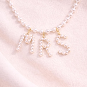May include: A delicate pearl necklace with the letters "MRS" spelled out in individual pearl clusters. The letters hang from a strand of pearls, creating a sophisticated and elegant accessory. The background is a soft, light pink fabric.