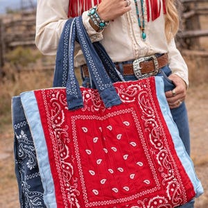 May include: A large tote bag with a red bandana print center, blue denim sides, and light blue trim. The bag has blue denim handles. The bag's dimensions are approximately 46 cm wide and 38 cm tall. The bag is held by a person wearing a western-style shirt and jeans.