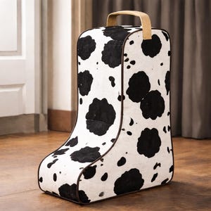 May include: A tall, boot-shaped storage unit with a cowhide pattern. The unit is white with large black spots and a brown trim. It has a tan handle at the top for easy carrying.