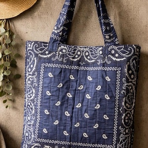 May include: A navy blue tote bag with a white paisley pattern. The bag is quilted and has matching handles. A straw hat hangs nearby. The bag is a stylish accessory for carrying items.