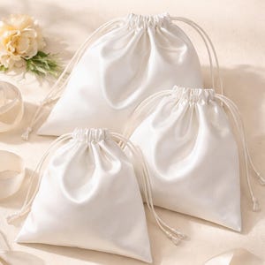 Puede incluir: Tres bolsas de viaje de satén blanco con cierres de cordón. El texto "Sewing Pattern" y "WHITE SATIN TRAVEL POUCHES" es visible. Una flor y una cinta están en el fondo. Las bolsas están hechas de satén brillante.