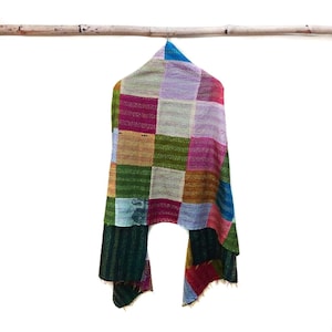 May include: A colorful patchwork scarf with a variety of square patterns in shades of green, pink, purple, and blue. The scarf is hanging from a bamboo rod, showcasing its vibrant design and texture.