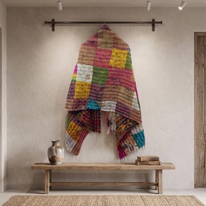 May include: A rectangular patchwork textile hanging from a dark metal rod, featuring a vibrant array of multicolored fabric squares in shades of yellow, pink, green, and blue with visible stitching patterns throughout the entire surface area.