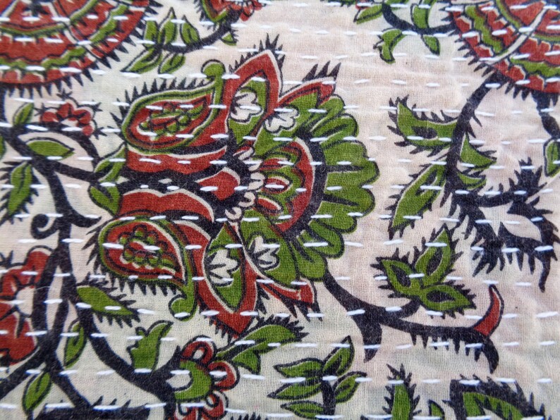 May include: A close-up of a white fabric with a floral pattern in shades of green, red, and black. The fabric is stitched with white thread, creating a quilted effect.