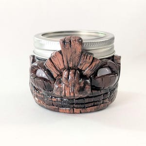 Pode incluir: Um suporte de jarro com tema tiki marrom e preto com tampa prateada. O suporte do jarro tem um design de rosto esculpido com olhos grandes e um sorriso largo. O suporte do jarro é feito de um material texturizado.