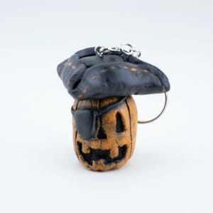 May include: A Halloween-themed keychain featuring a carved pumpkin with a pirate hat and eye patch. The pumpkin is orange with a carved face, and the hat is black with a silver keychain ring.