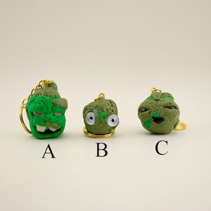 May include: Three textured, green, sculpted character keychains labeled A, B, and C. Keychain A is a bright green head with large white teeth. Keychain B is a round, olive-green shape with two purple eyes. Keychain C is a round, olive-green head.