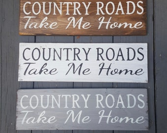 May include: Three wooden signs with the text "COUNTRY ROADS Take Me Home" painted in white, black, and gray. The signs are made of rustic wood and have a distressed look.