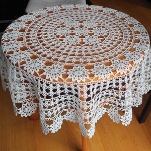 Op de afbeelding: Een rond wit gehaakt tafelkleed met een ingewikkeld bloemen- en geometrisch patroon met opengewerkte details. De kanten stof valt over de randen van een ronde houten tafel en toont een herhalend stervormig motief en een geschulpte rand.