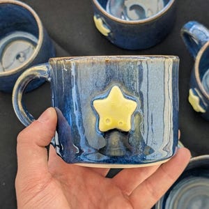 May include: Handmade ceramic mug with a blue glaze and a yellow star applique. The mug has a rounded handle and a slightly textured surface. Other similar mugs are visible in the background. The mug is approximately 9 cm tall.