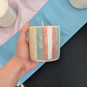 Puede incluir: Una vela cilíndrica con rayas verticales en colores pastel beige, azul, rosa y blanco. La vela se sostiene en una mano, y el fondo incluye una tela azul claro y rosa.