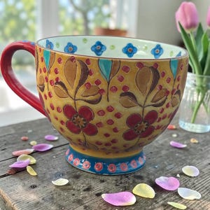 Pu&ograve; includere: Una grande tazza decorativa in ceramica con manico e base rossi. La tazza presenta uno sfondo giallo con motivi floreali marroni, rossi e turchesi. Il bordo interno presenta disegni di fiori blu. Petali sparsi sulla superficie di legno.